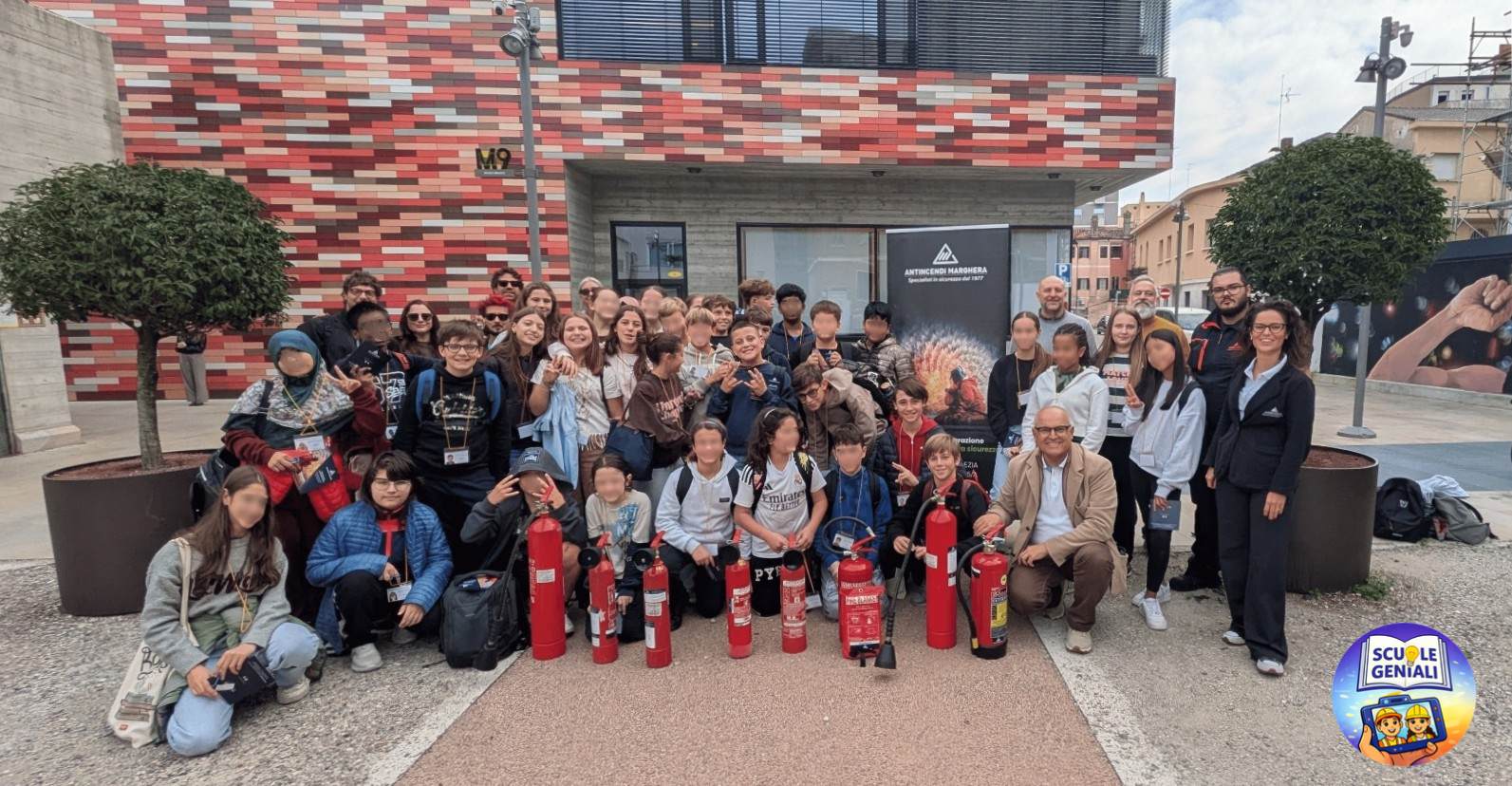 Gruppo di studenti e adulti in posa davanti a un edificio, con estintori in primo piano. L'immagine mostra un ambiente urbano con alberi e pareti colorate sullo sfondo.