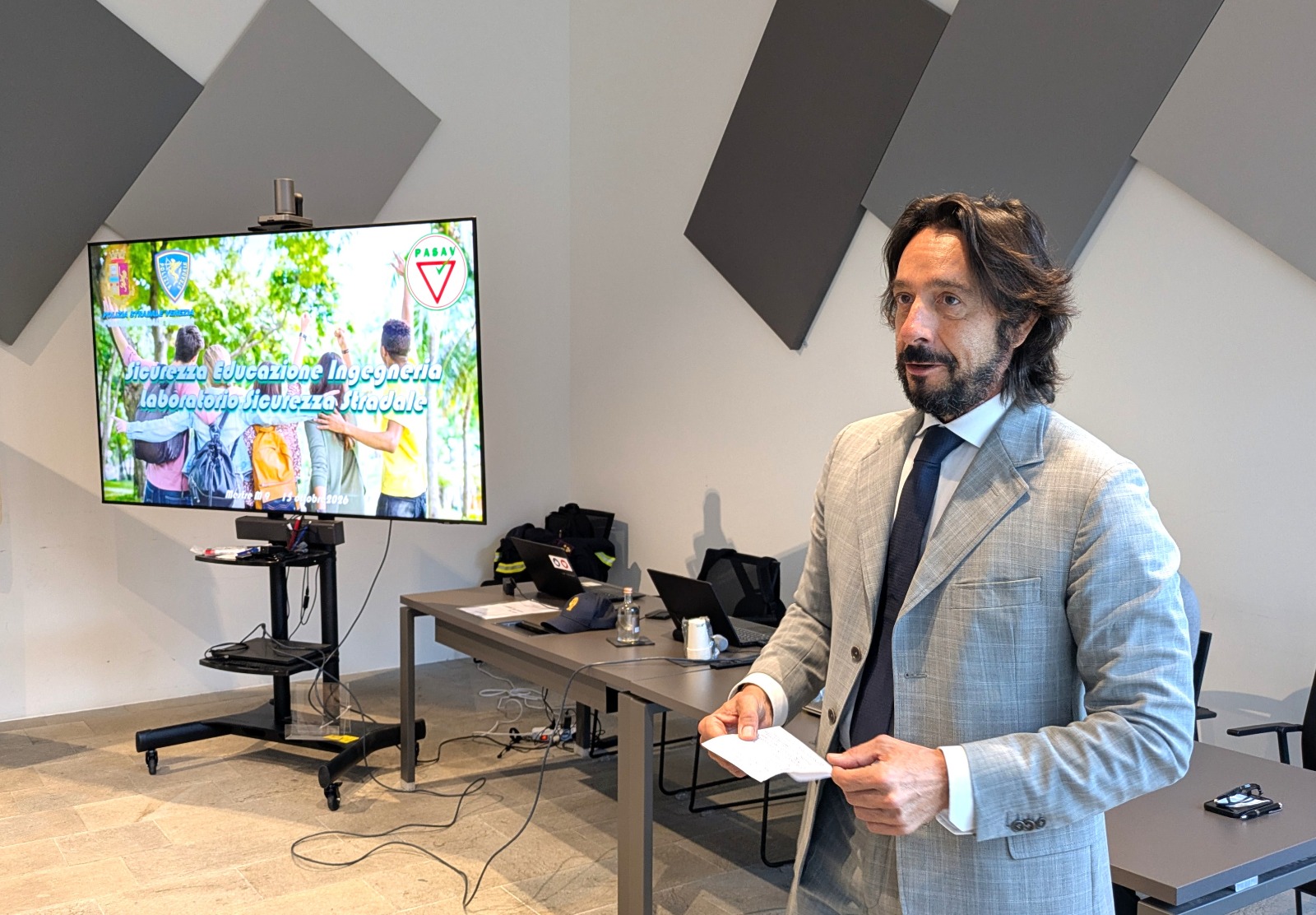 Un presentatore in un abito chiaro parla di fronte a uno schermo che mostra informazioni sulla sicurezza stradale e l'ingegneria, con un laboratorio in primo piano.