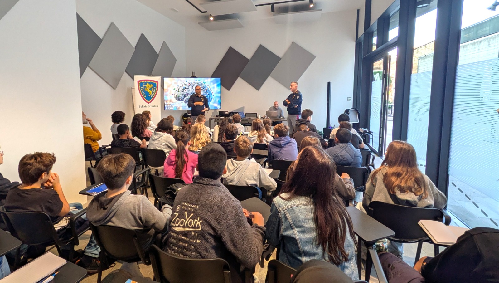 Un gruppo di studenti ascolta una presentazione in una sala moderna. Due relatori, uno dei quali è un membro della Polizia Stradale, discutono di un tema legato alla sicurezza, mentre un altro uomo è seduto a un tavolo con un computer. Sullo sfondo c'è uno schermo con un'immagine.