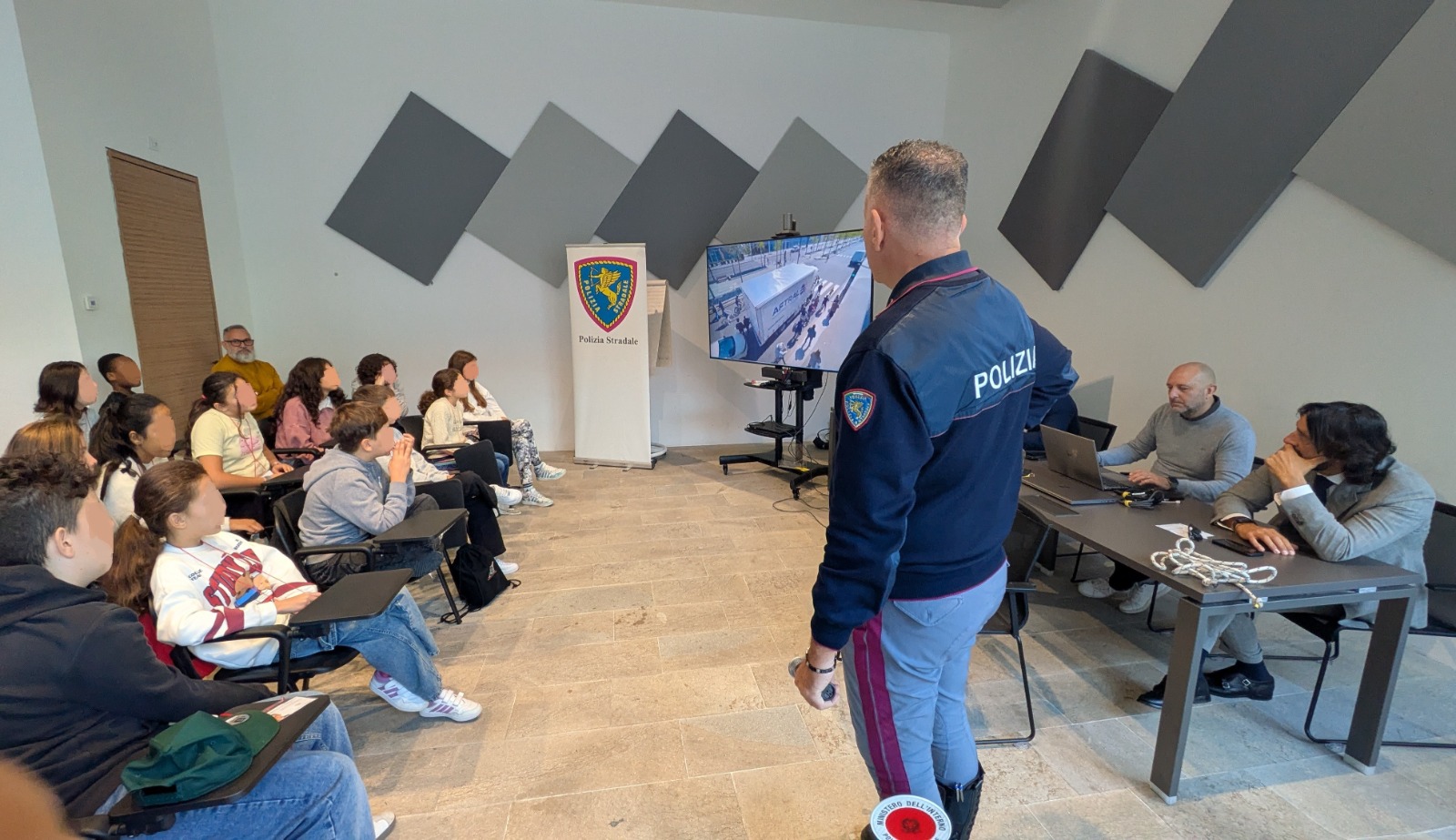Un agente di polizia sta presentando davanti a un gruppo di giovani, con un video proiettato su uno schermo. Gli studenti siedono in modo attento mentre ascoltano la presentazione all'interno di una sala.
