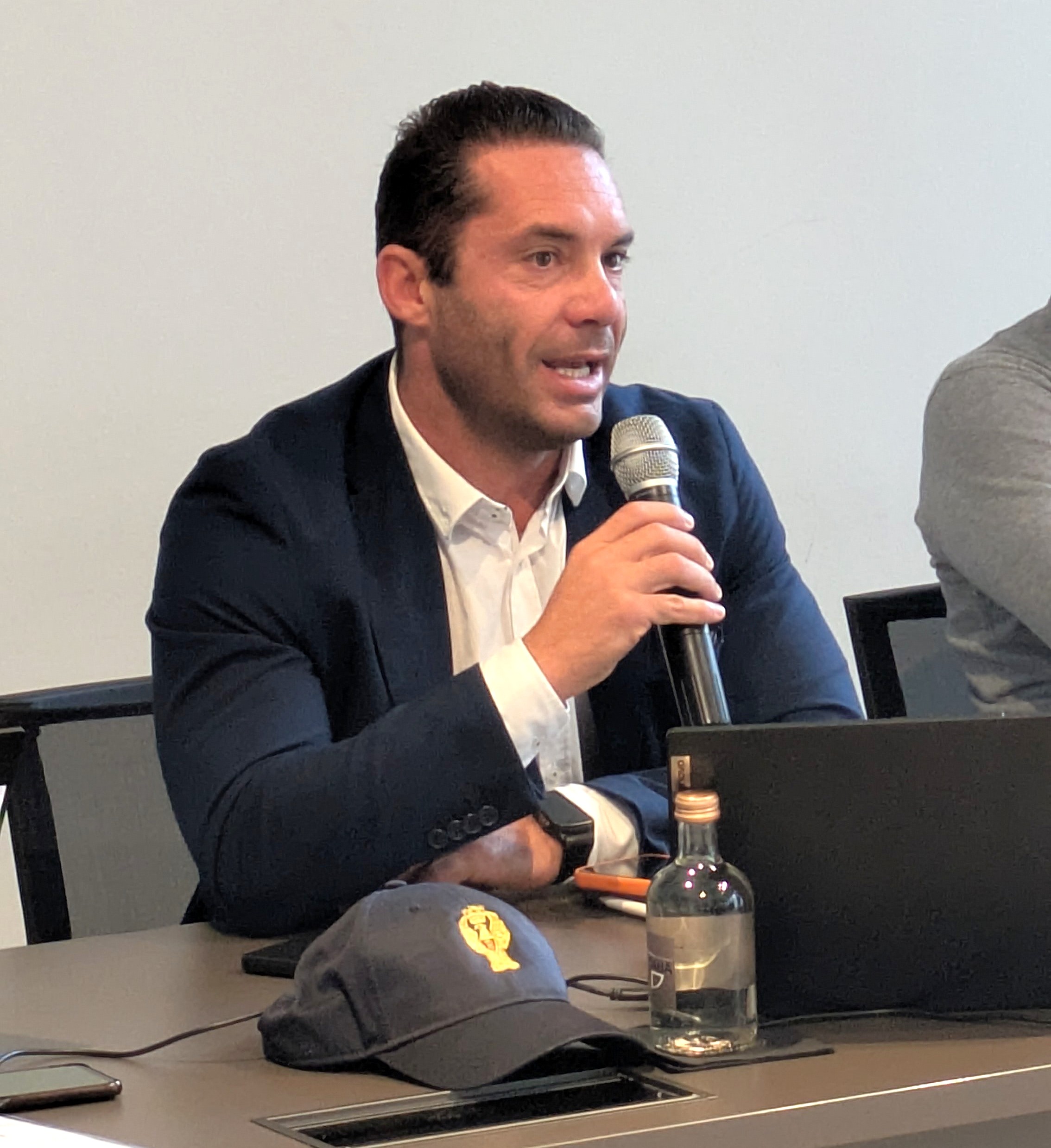 Uomo con giacca scura e camicia bianca che parla al microfono durante una conferenza, con un computer portatile e una bottiglia d'acqua sul tavolo.