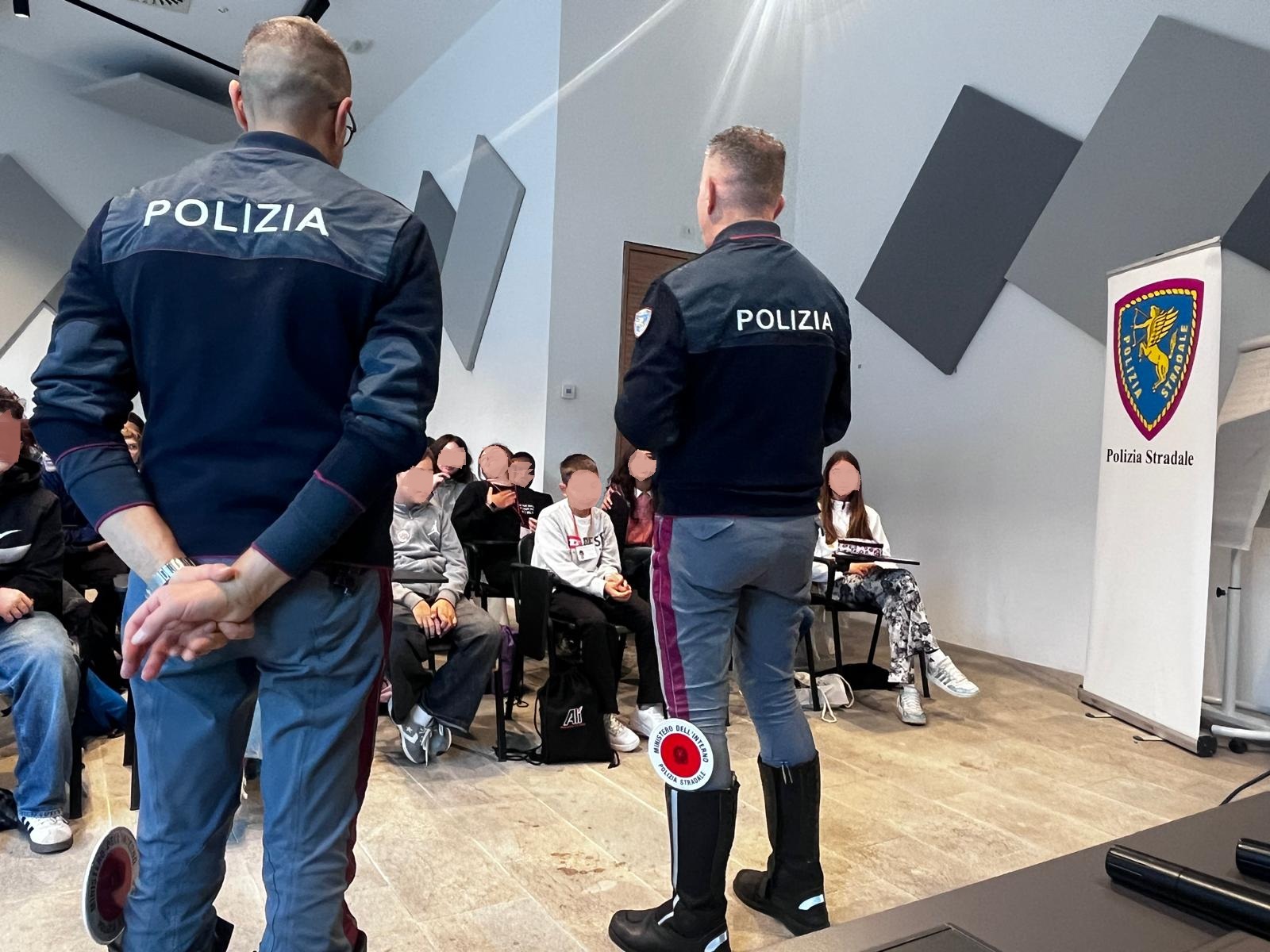 Due agenti della polizia in uniforme si rivolgono a un gruppo di giovani in una sala, mentre un banner della Polizia Stradale è visibile sullo sfondo.