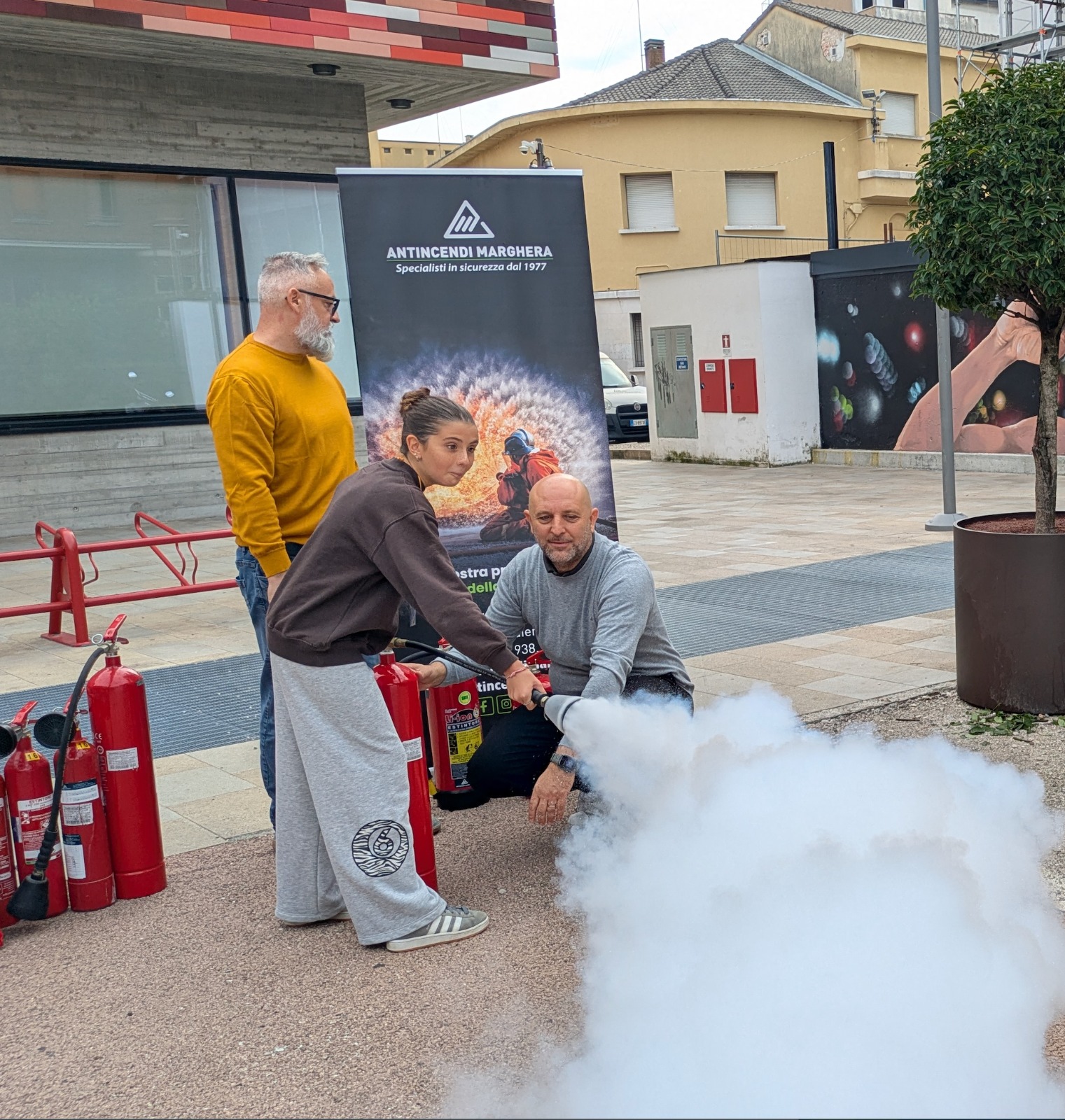 Una donna utilizza un estintore mentre un uomo le mostra la tecnica corretta, in un'area pubblica con estintori visibili e un banner di sicurezza sullo sfondo.